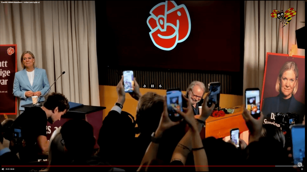 Konferencja prasowa z liderami, z jedną osobą na podium w niebieskim garniturze i tłumem trzymającym telefony, zdjęcie z tłem z logo partii.