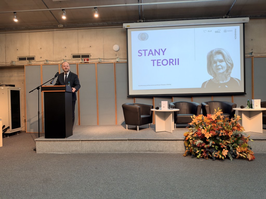 Prezentacja na konferencji 'Stany Teorii' z mówcą stojącym przy mównicy, z ekranem w tle przedstawiającym nazwę konferencji i zdjęcie prelegentki.