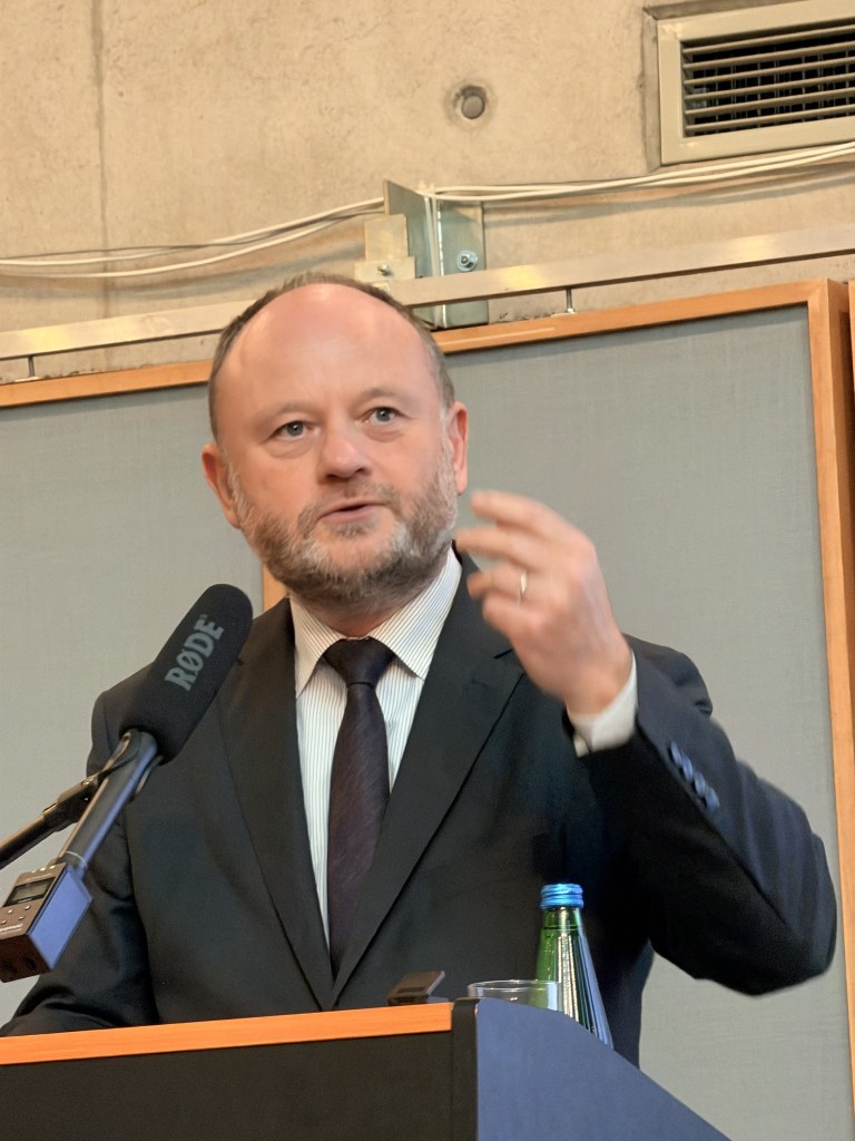 Mężczyzna w garniturze przemawiający na konferencji, z mikrofonem i prezentacją w tle.