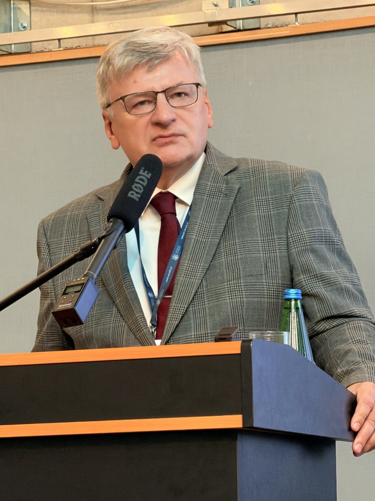 Mężczyzna w szarej, w kratkę marynarce stoi przy mównicy, trzyma mikrofon i wygląda na zamyślonego.
