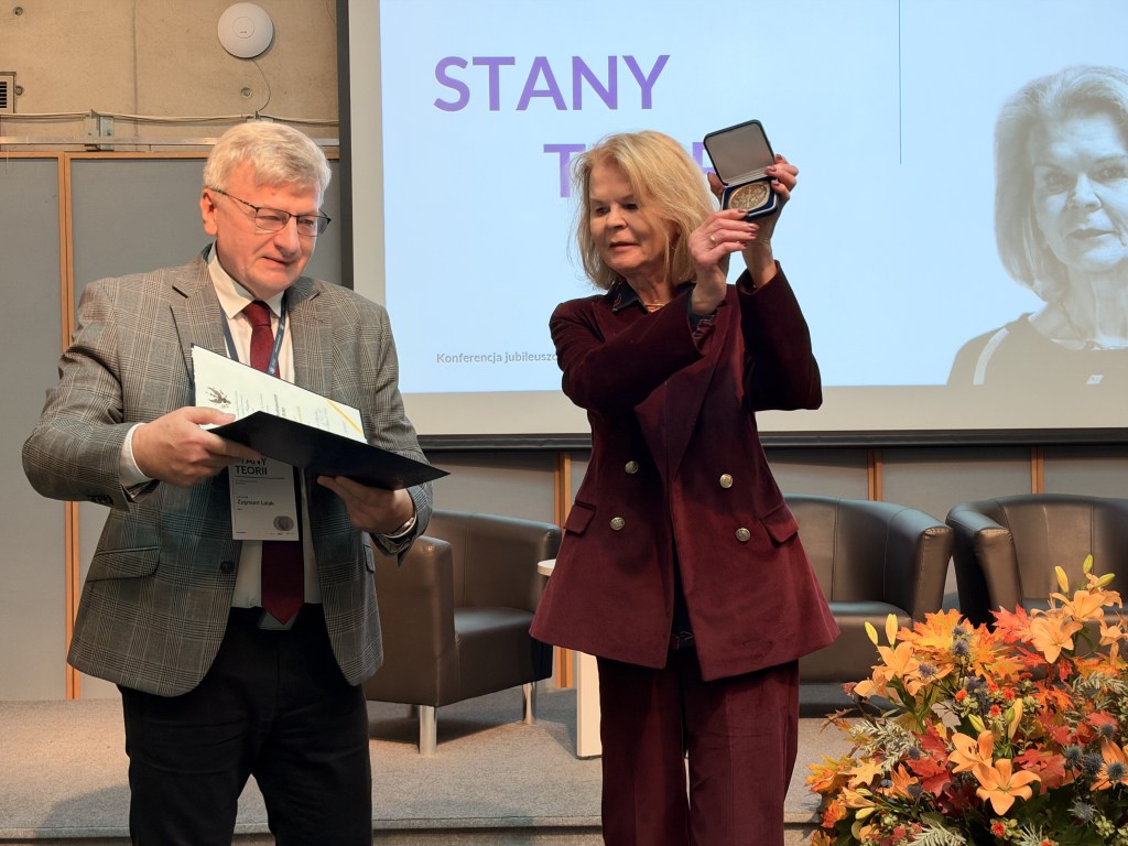 Uczestnicy konferencji wręczają nagrodę, jeden z mężczyzn trzyma dokument, a kobieta prezentuje medal na scenie.