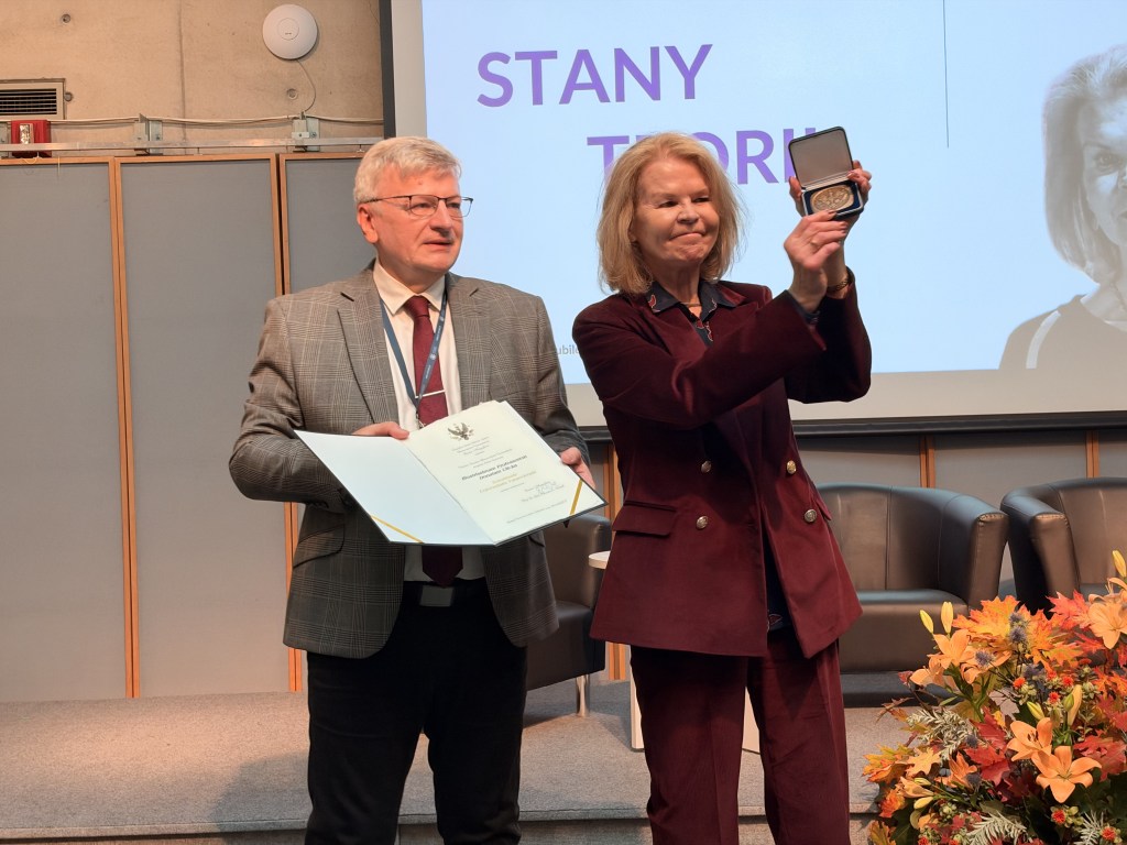 Mężczyzna w garniturze trzyma dyplom, a kobieta w ciemnej sukience uśmiecha się, trzymając medal w dłoni. W tle widoczne fotele oraz kwiaty.