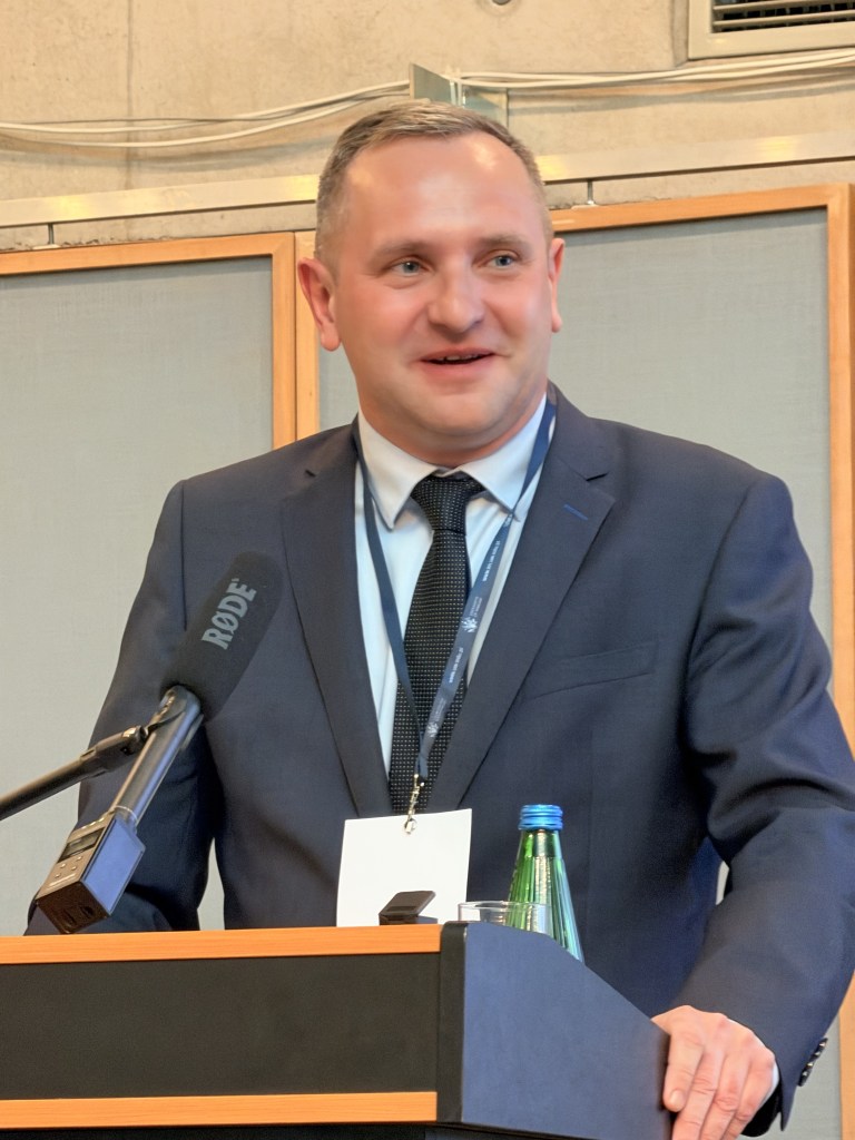 Mężczyzna w garniturze z uśmiechem, przemawiający z mównicy podczas wydarzenia. W tle widoczne elementy sali konferencyjnej.
