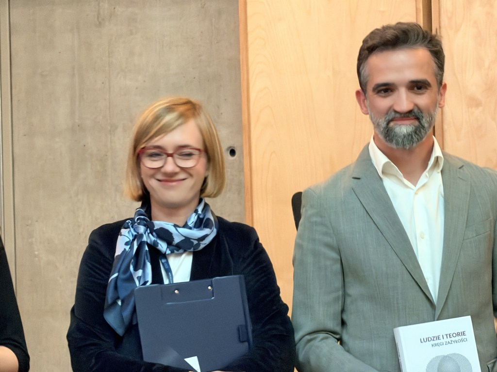 Dwoje uśmiechniętych ludzi stojących obok siebie podczas wydarzenia, na tle betonowej ściany.