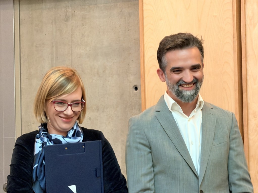Dwoje uśmiechniętych ludzi stojących obok siebie na tle szarej i drewnianej ściany. Kobieta z blond włosami i okularami, trzymająca teczkę, oraz mężczyzna z siwą brodą w szarym garniturze.