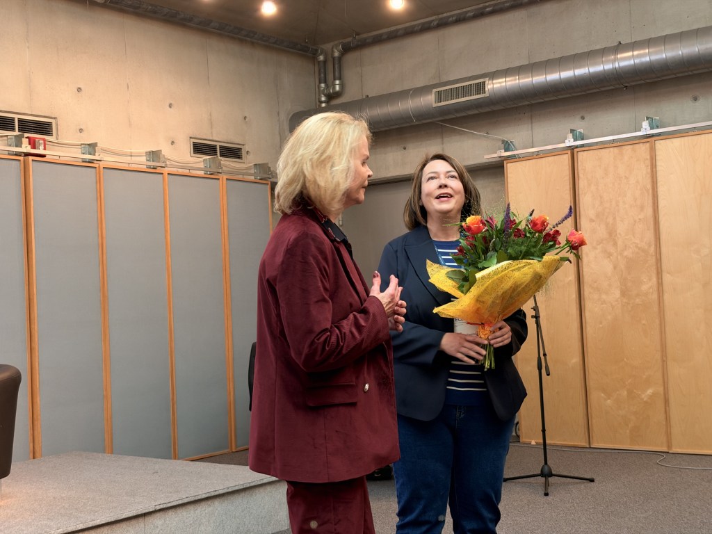 Dwie kobiety rozmawiają na tle kwiatów, jedna z nich trzyma bukiet w dłoniach, a druga słucha z uśmiechem.