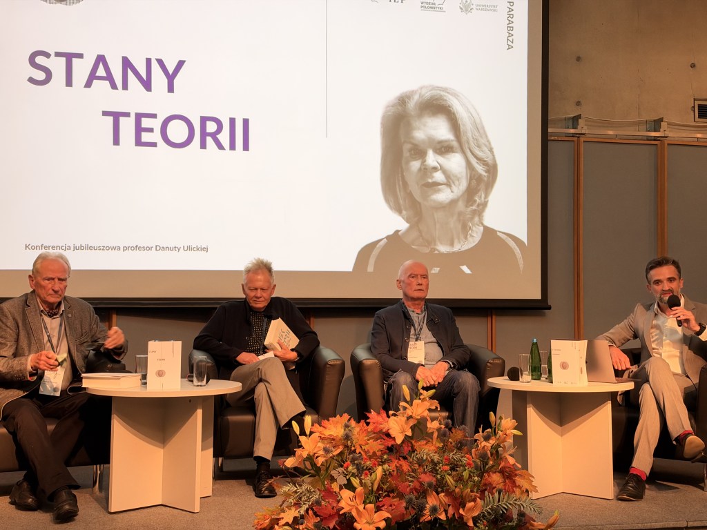Czterech mężczyzn siedzących na scenie podczas konferencji z okazji jubileuszu profesor Danuty Ulickiej. W tle widoczny portret profesor Ulickiej oraz napis 'STANY TEORII'.