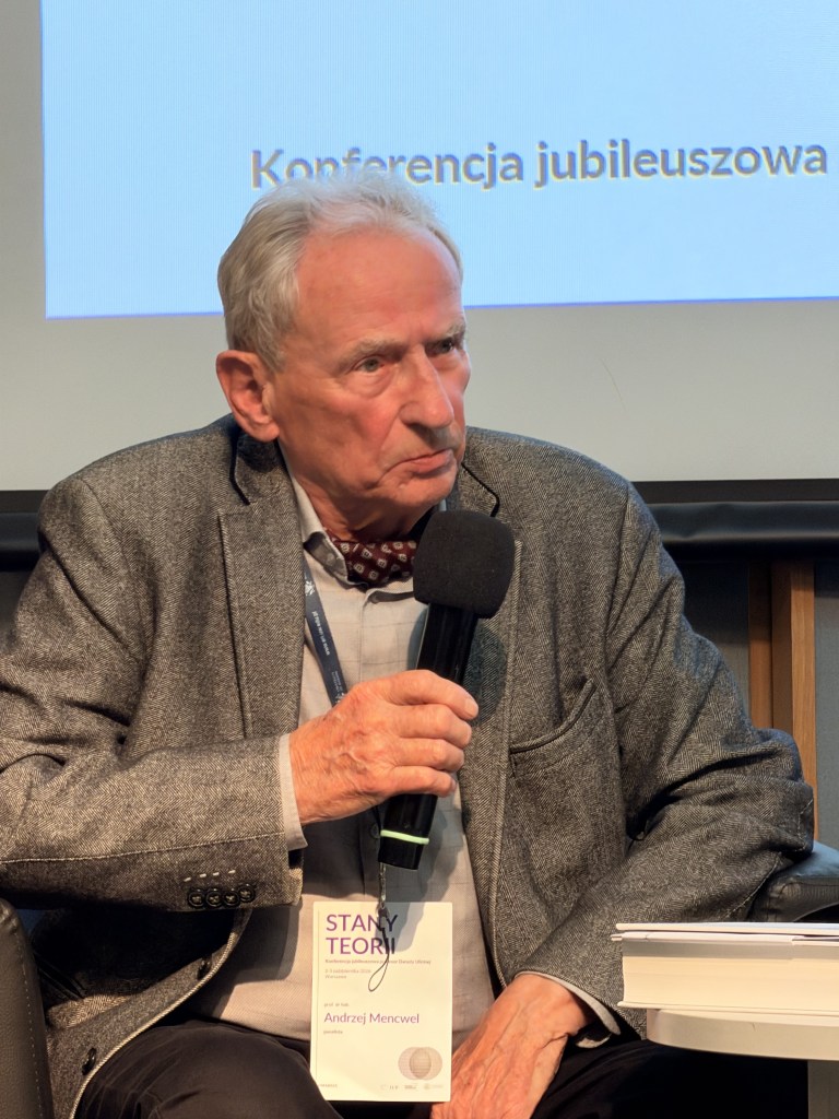 Mężczyzna z szarym garniturem siedzi na scenie z mikrofonem, uczestnicząc w konferencji jubileuszowej.