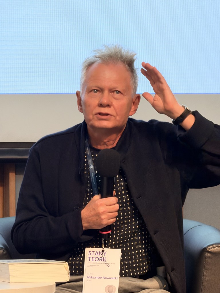 Mężczyzna trzyma mikrofon i gestykuluje podczas wystąpienia, siedząc na panelu dyskusyjnym. W tle projektor wyświetla nieczytelny tekst.