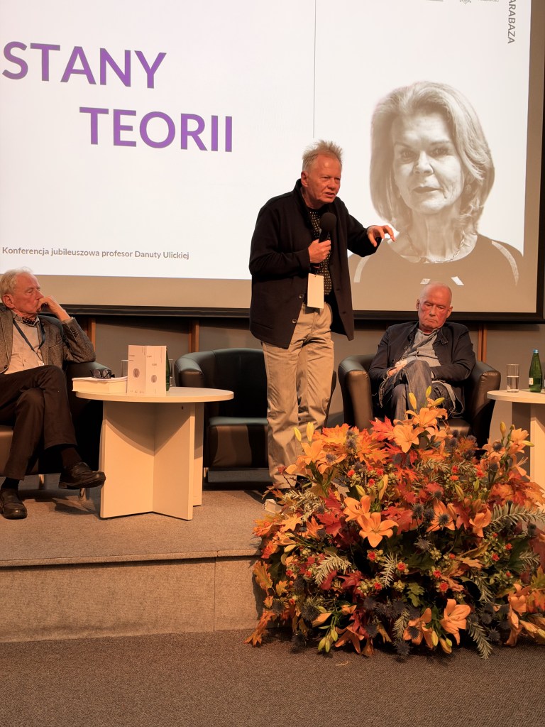 Konferencja jubileuszowa z okazji urodzin profesor Danuty Ulickiej, na której przemawia jeden z panelistów, przy dwóch siedzących uczestnikach i kwiatowej dekoracji na pierwszym planie.