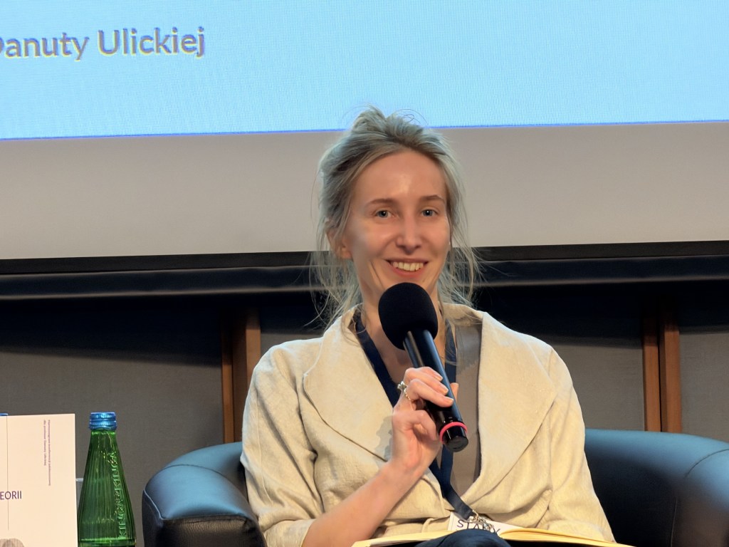 Kobieta z mikrofonem, uśmiechająca się, siedząca w czarnej sofie podczas wydarzenia, z ekranem w tle.