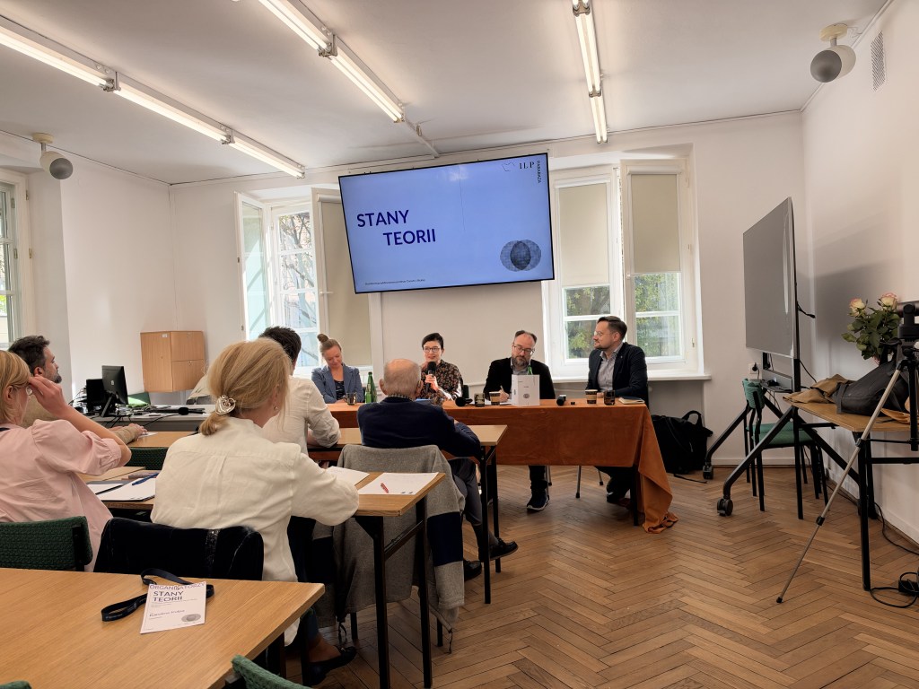 Spotkanie dyskusyjne na temat "Stany Teorii" z prelegentami siedzącymi za stołem, w sali z oknami i rzędem uczestników siedzących na krzesłach.