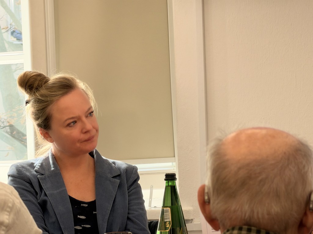 Kobieta słuchająca w dyskusji, z blond włosami związanymi w kok, w niebieskim żakiecie, w tle mężczyzna z baldą głową.