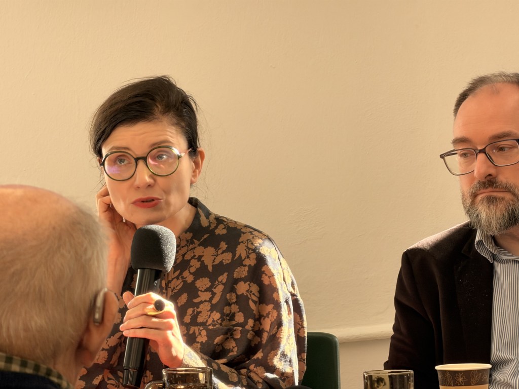 Kobieta z mównicą trzyma mikrofon, mówi do publiczności, obok siedzi mężczyzna w okularach.