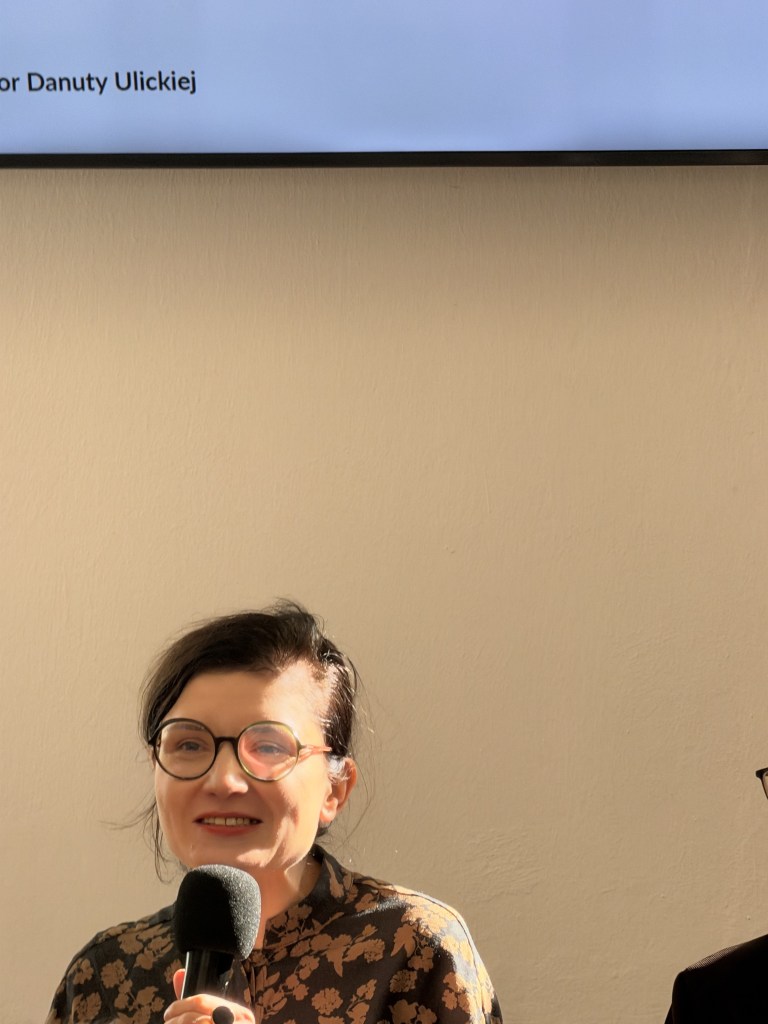 Kobieta z okularami, trzymająca mikrofon podczas wydarzenia, uśmiecha się do kamery.